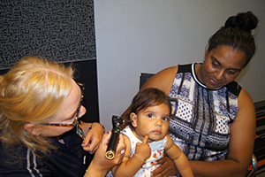 A health professional looks into a toddler's ear. The toddler is seated on woman's lap.