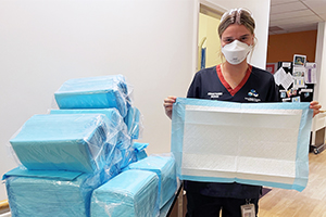 A female nurse wearing a surgical mask holds a blue absorbant pad known as a bluey. She stands beside a trolley stacked with blueys.