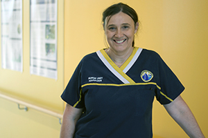 A woman wearing a Rockingham General Hospital uniform that reads Clinical nurse consultant stands in front of a coloured wall.