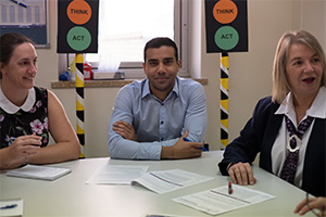 A man and two women sit at a table in conversation. Behind the man are two displays that look like traffic lights. On the amber light is the word 'Think' while on the green light is the word 'Act'.