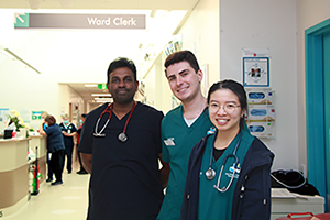 Three doctors (two male and one female) stand in a hospital corridor. Behind them in the corridor is a sign that reads Ward Clerk