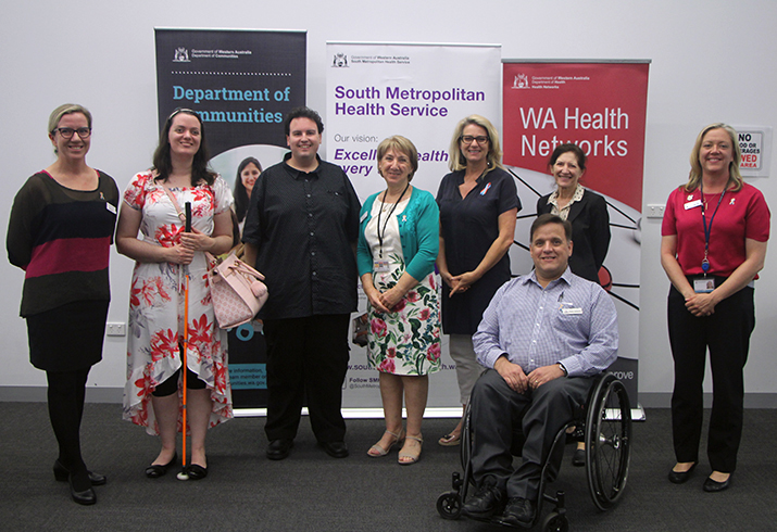 A group of eight people in front of three banners that read 'Department of Communities', 'South Metropolitan Health Service' and 'WA Health Networks'. One woman is holding a cane and one man is in a wheelchair.
