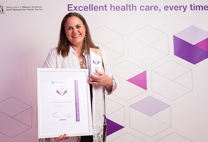 Dr Vanessa Clayden holds a SMHS Excellence Awards certificate and trophy. Behind her a banner reads excellent health care, every time.