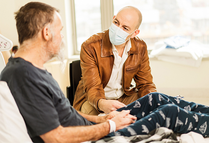 A man wearing a surgical mark sits talking to a man sitting in a hospital bed.