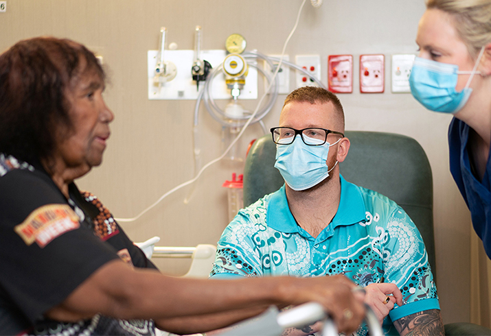 A male and female health professional talk with an Aboriginal woman. The woman is sitting down and her hands are resting on a walking frame.