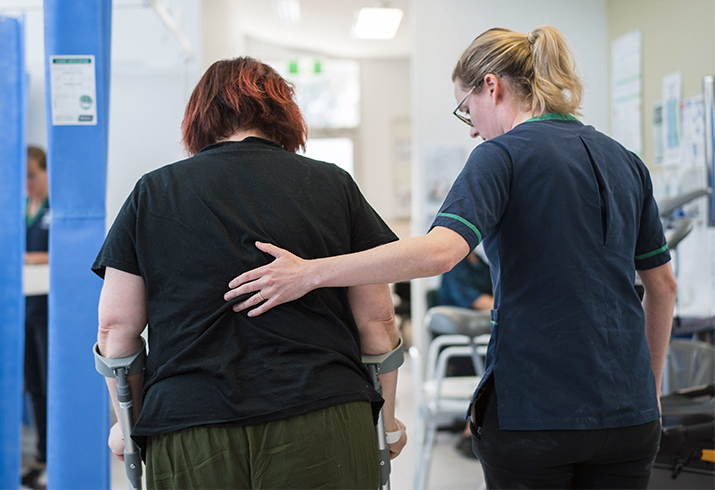 A female therapist places a supportive hand on the back of a female patient using elbow crutches