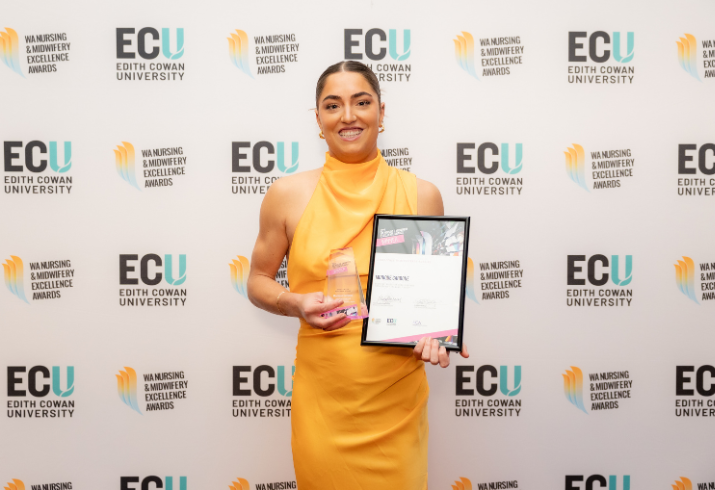 Monique standing in a yellow dress, holding a framed certificate and glass trophy.