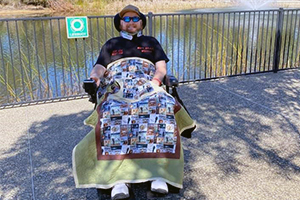 A man in a wheelchair with a blanket over his legs sits in front of a lake