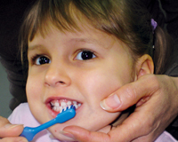 Woman holding a young girl's chin from behind while brushing the girl's bottom teeth.